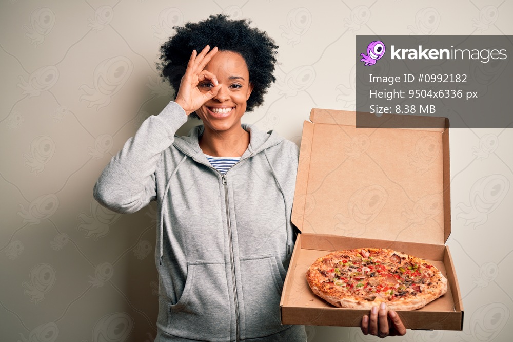 Young African American afro woman with curly hair holding delivery box with Italian pizza with happy face smiling doing ok sign with hand on eye looking through fingers