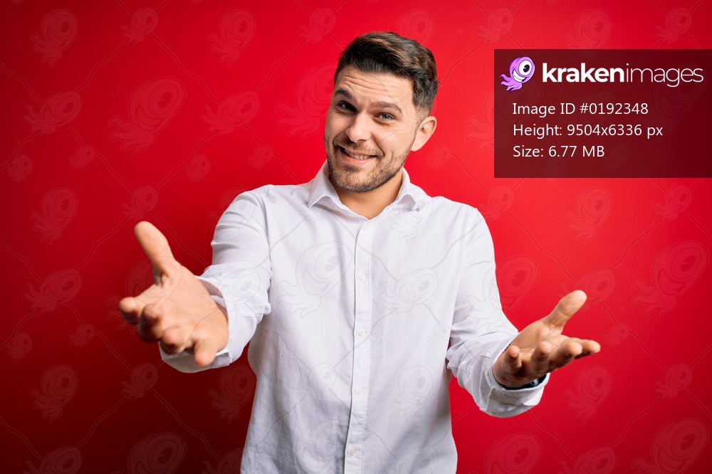 Young business man with blue eyes wearing elegant shirt standing over red isolated background smiling cheerful with open arms as friendly welcome, positive and confident greetings