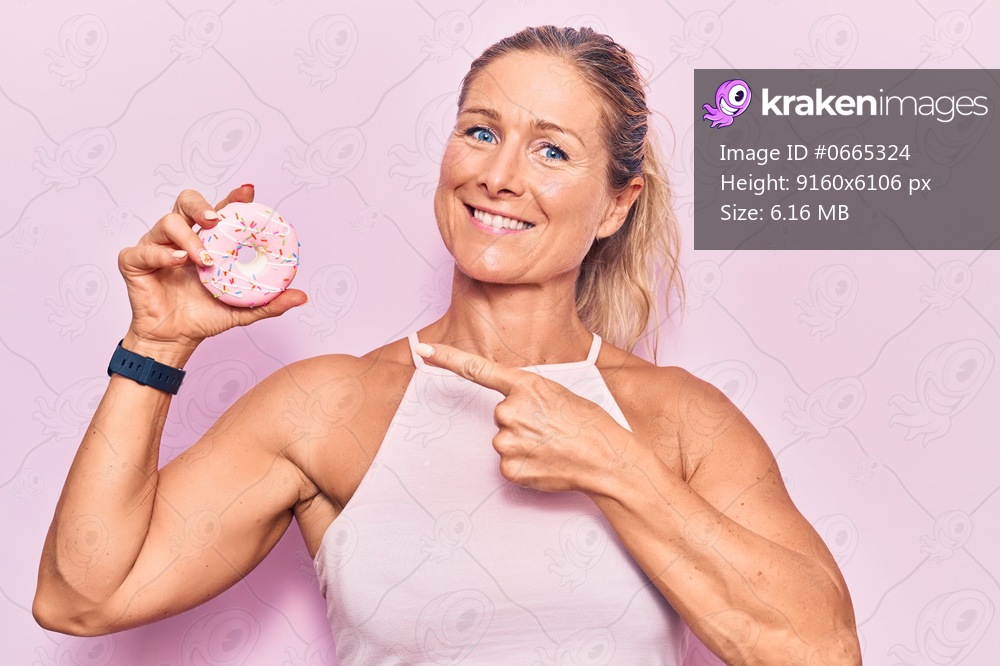 Middle age caucasian blonde woman wearing sporty clothes and holding sugar doughnut smiling happy pointing with hand and finger 