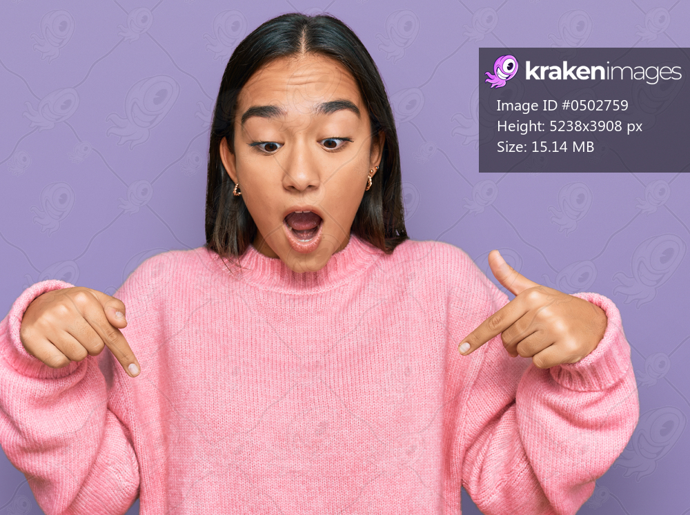 Young asian woman wearing casual winter sweater pointing down with fingers showing advertisement, surprised face and open mouth 