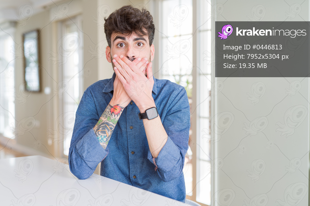 Young man wearing casual shirt sitting on white table shocked covering mouth with hands for mistake. Secret concept.