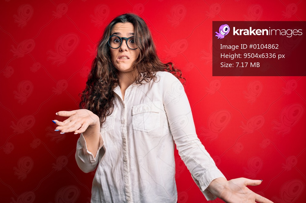 Young beautiful woman with curly hair wearing shirt and glasses over red background clueless and confused expression with arms and hands raised. Doubt concept.