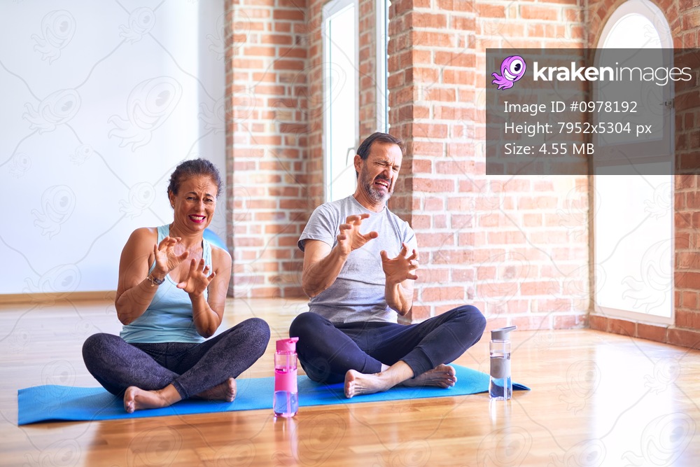 Middle age sporty couple sitting on mat doing stretching yoga exercise at gym disgusted expression, displeased and fearful doing disgust face because aversion reaction. With hands raised. Annoying concept.