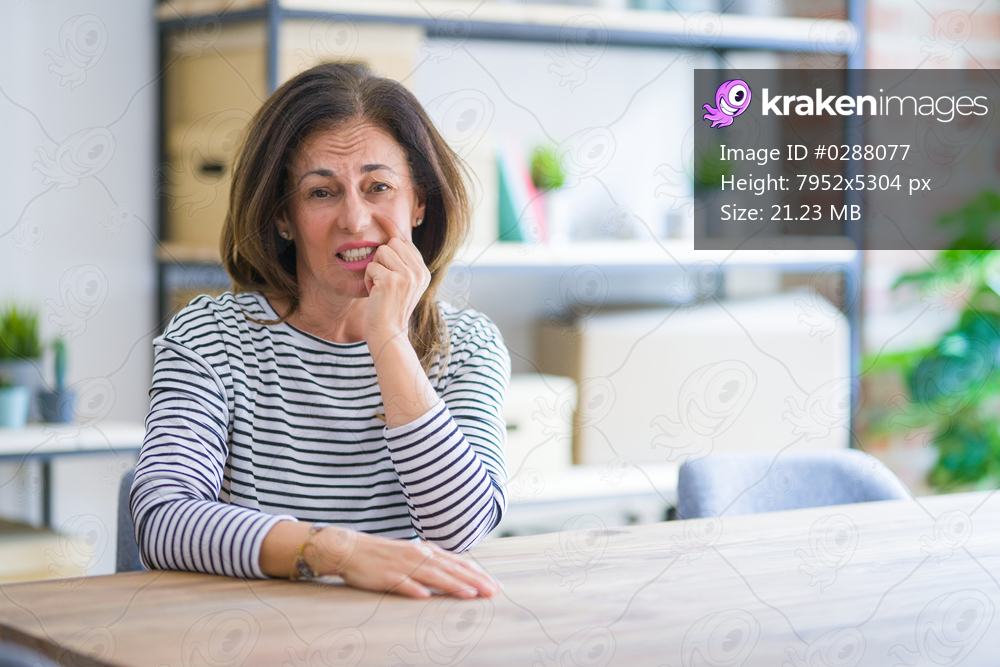 Middle age senior woman sitting at the table at home looking stressed and nervous with hands on mouth biting nails. Anxiety problem.