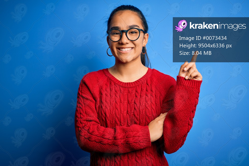 Young beautiful asian woman wearing casual sweater and glasses over blue background with a big smile on face, pointing with hand and finger to the side looking at the camera.