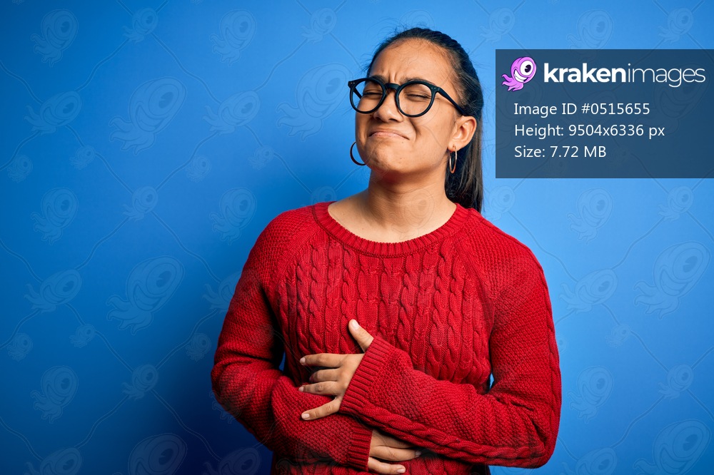 Young beautiful asian woman wearing casual sweater and glasses over blue background with hand on stomach because nausea, painful disease feeling unwell. Ache concept.