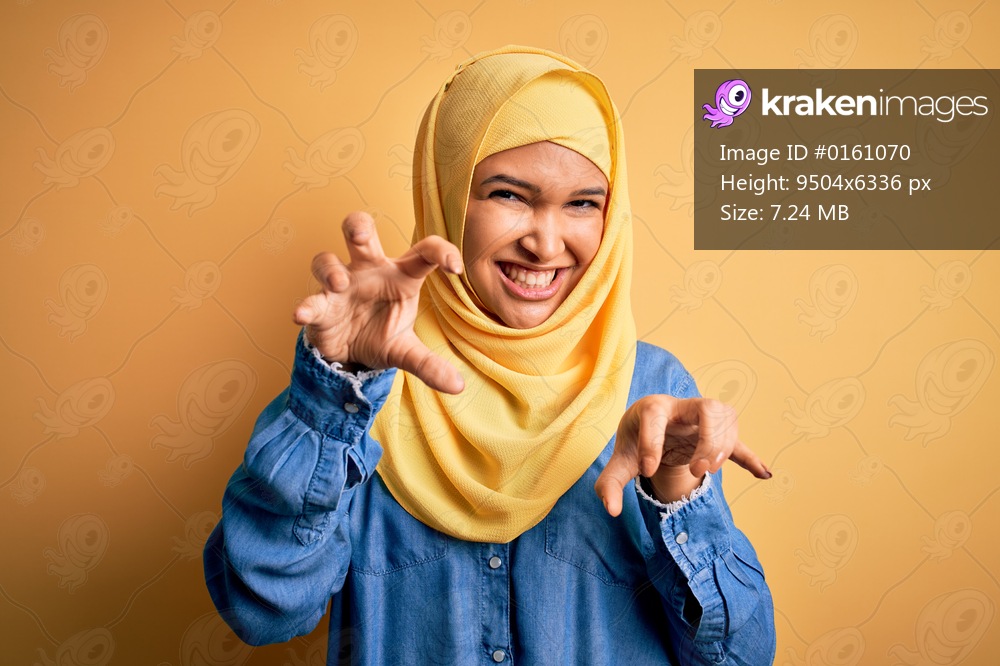 Young beautiful woman with curly hair wearing arab traditional hijab over yellow background smiling funny doing claw gesture as cat, aggressive and sexy expression