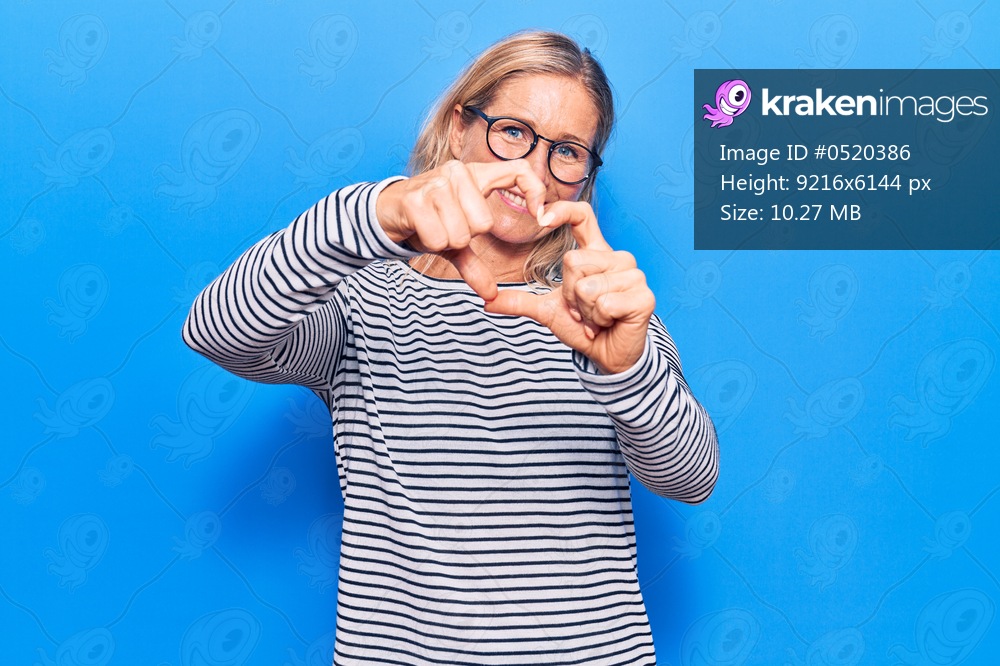 Middle age caucasian blonde woman wearing casual striped sweater and glasses smiling in love doing heart symbol shape with hands. romantic concept. 