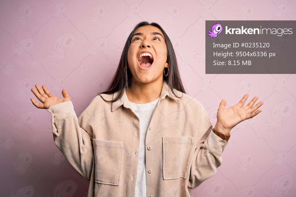 Young beautiful asian woman wearing casual shirt standing over pink background crazy and mad shouting and yelling with aggressive expression and arms raised. Frustration concept.