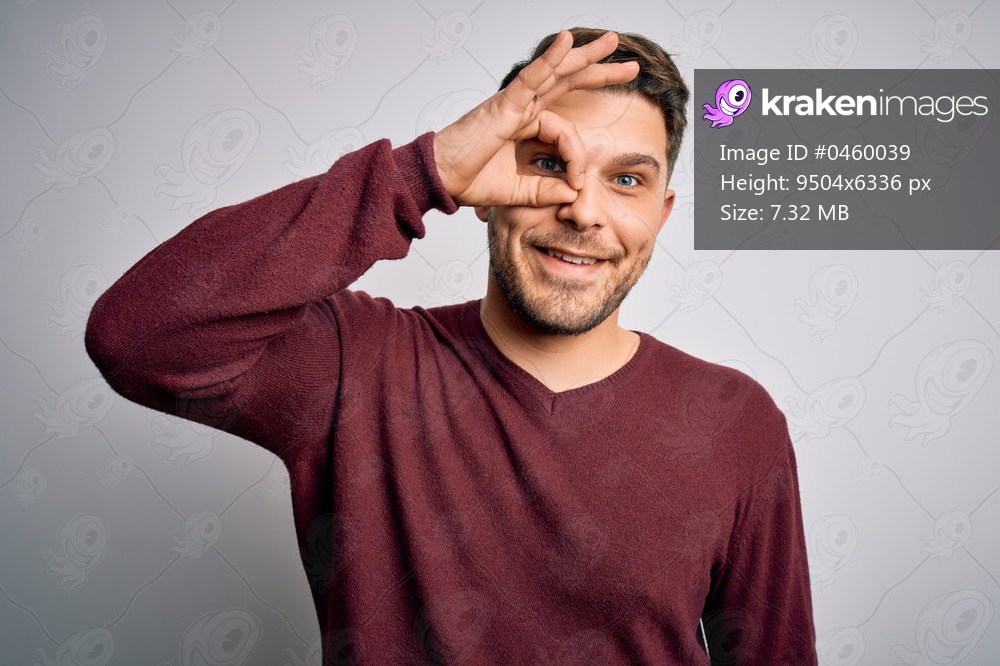 Young man with blue eyes wearing casual sweater standing over isolated background doing ok gesture with hand smiling, eye looking through fingers with happy face.