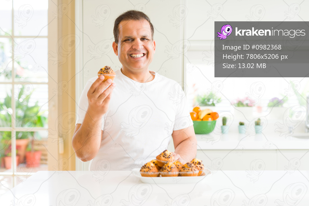 Middle age man eating chocolate chip muffins at home with a happy face standing and smiling with a confident smile showing teeth