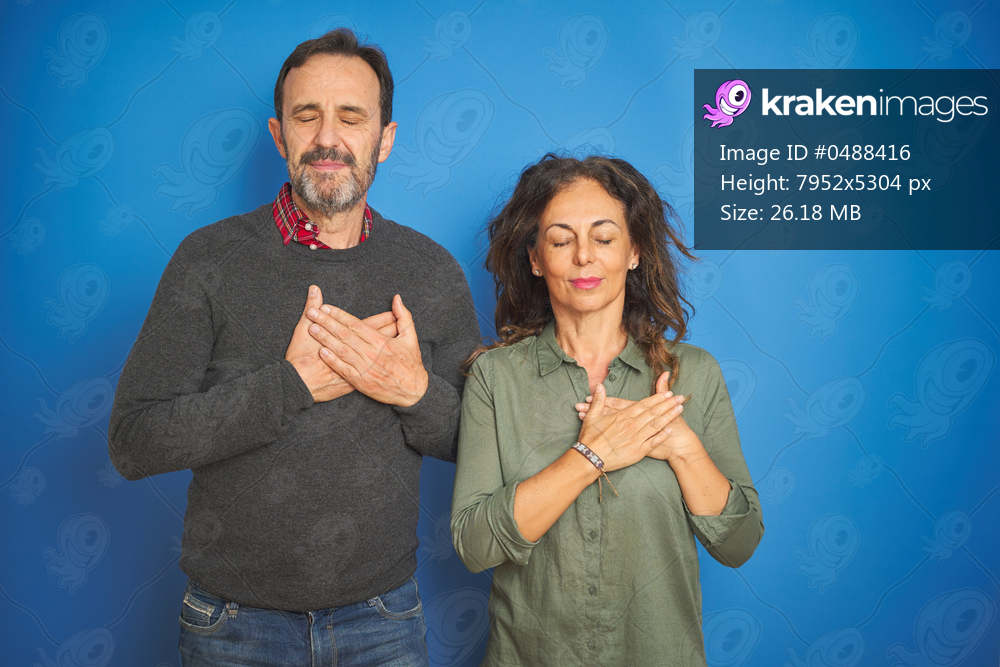 Beautiful middle age couple together standing over isolated blue background smiling with hands on chest with closed eyes and grateful gesture on face. Health concept.