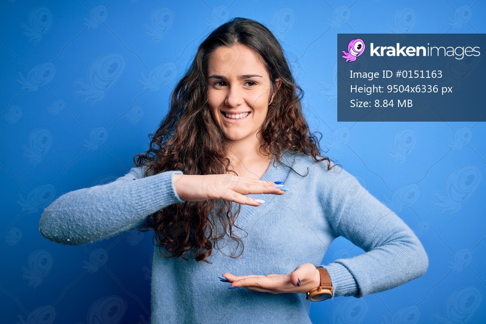 Young beautiful woman with curly hair wearing blue casual sweater over isolated background gesturing with hands showing big and large size sign, measure symbol. Smiling looking at the camera. Measuring concept.