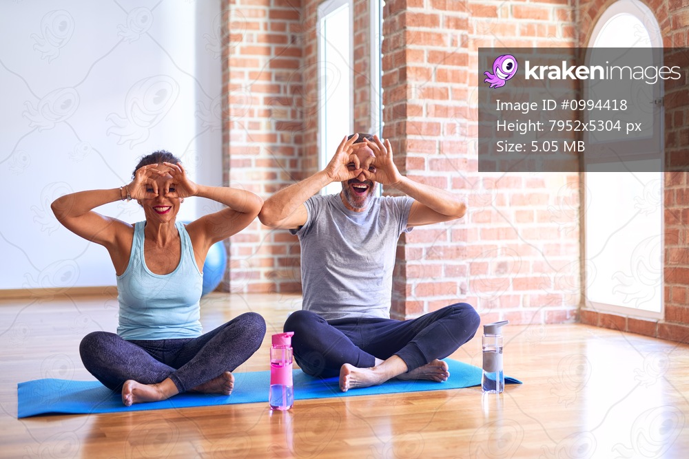 Middle age sporty couple sitting on mat doing stretching yoga exercise at gym doing ok gesture like binoculars sticking tongue out, eyes looking through fingers. Crazy expression.