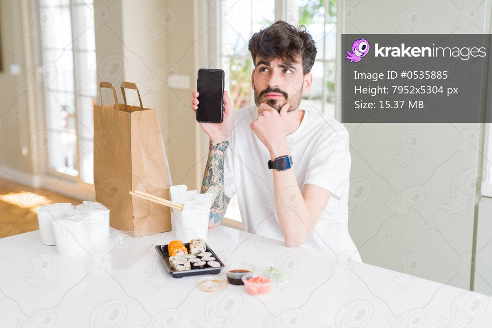Young man eating asian sushi from home delivery and showing smartphone screen serious face thinking about question, very confused idea