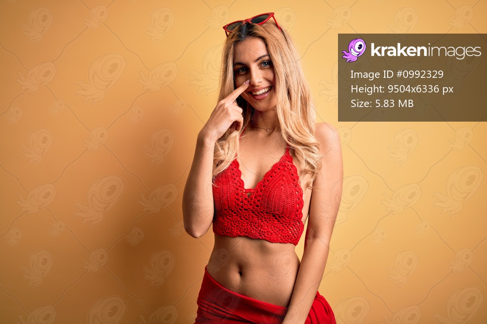 Young beautiful blonde woman on vacation wearing bikini over isolated yellow background Pointing with hand finger to face and nose, smiling cheerful. Beauty concept