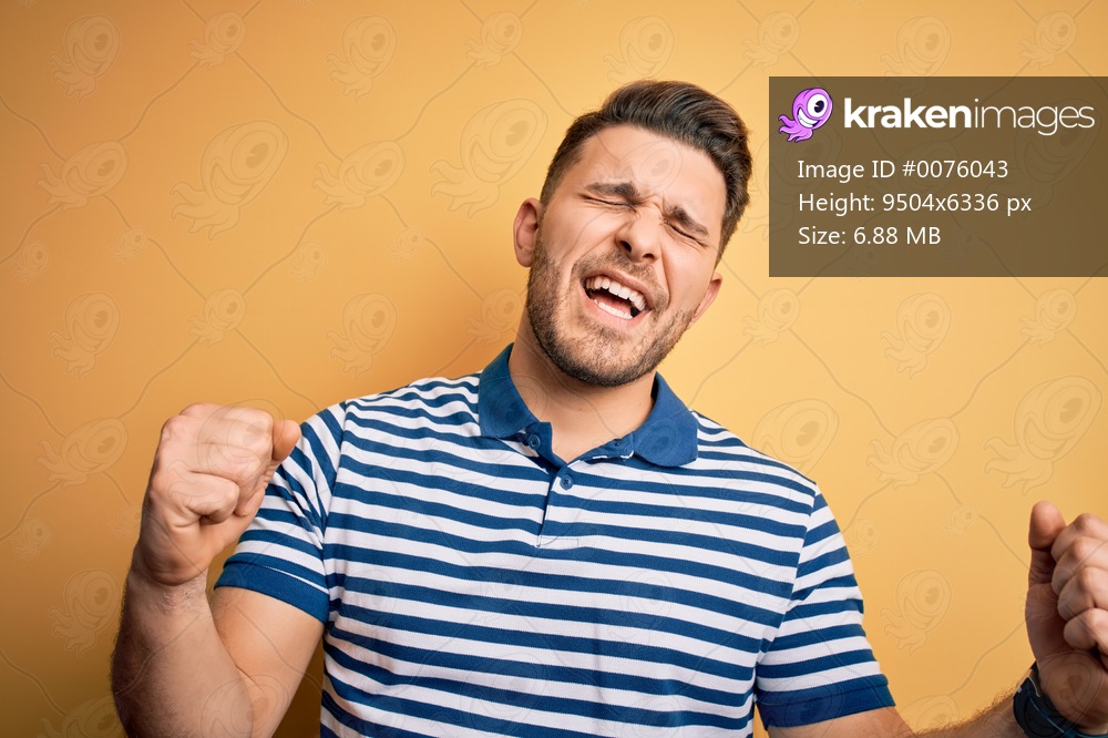 Young man with blue eyes wearing casual striped t-shirt over yellow background very happy and excited doing winner gesture with arms raised, smiling and screaming for success. Celebration concept.