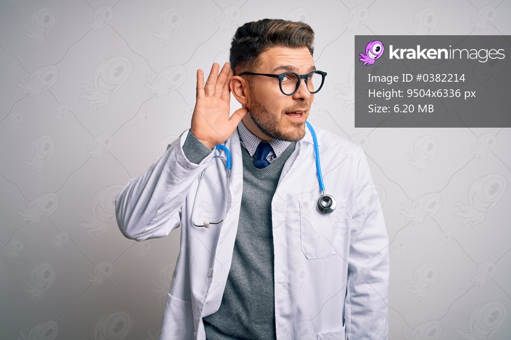 Young doctor man with blue eyes wearing medical coat and stethoscope over isolated background smiling with hand over ear listening an hearing to rumor or gossip. Deafness concept.