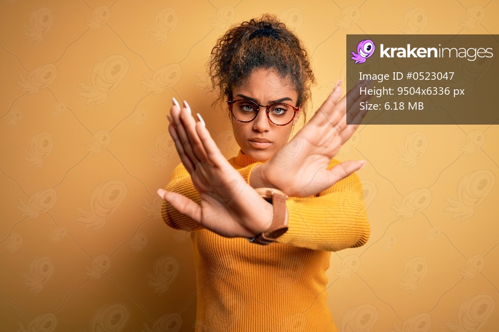 Young beautiful african american girl wearing sweater and glasses over yellow background Rejection expression crossing arms and palms doing negative sign, angry face