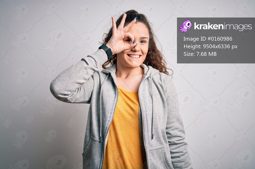 Young beautiful sportswoman with curly hair wearing sportswear over white background doing ok gesture with hand smiling, eye looking through fingers with happy face.
