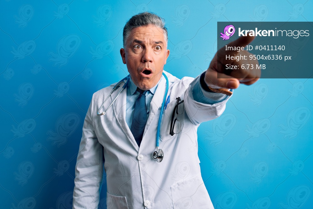 Middle age handsome grey-haired doctor man wearing coat and blue stethoscope Pointing with finger surprised ahead, open mouth amazed expression, something on the front