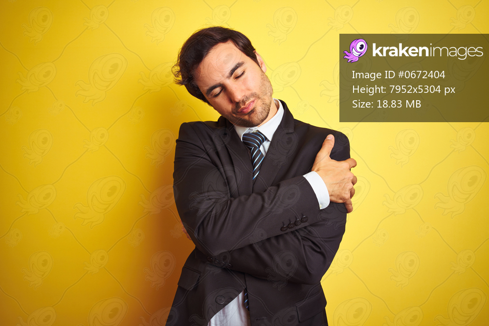 Young handsome businessman wearing suit and tie standing over isolated yellow background Hugging oneself happy and positive, smiling confident. Self love and self care