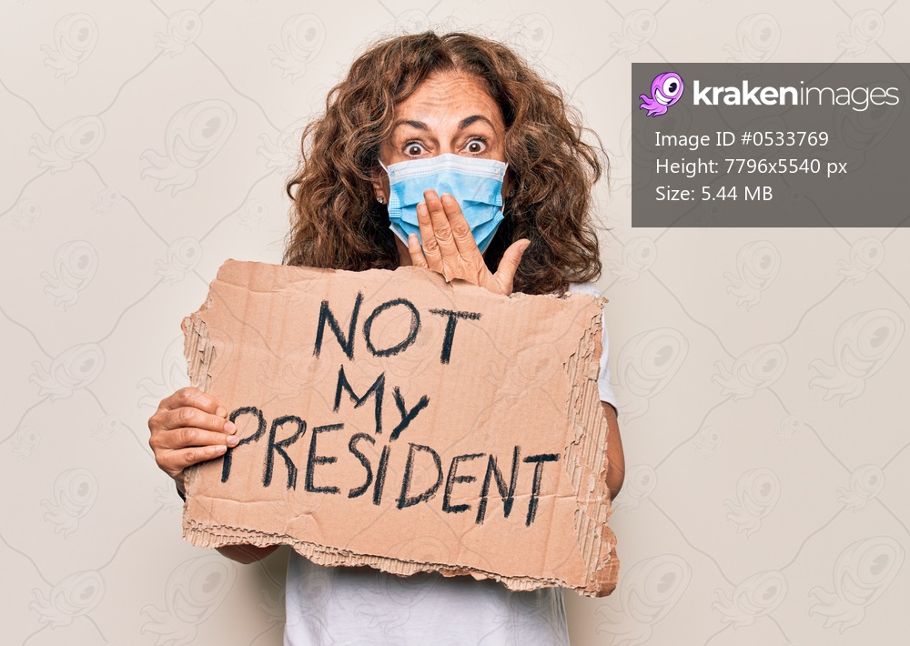 Middle age woman wearing coronavirus protection mask holding not my president protest cardboard covering mouth with hand, shocked and afraid for mistake. Surprised expression