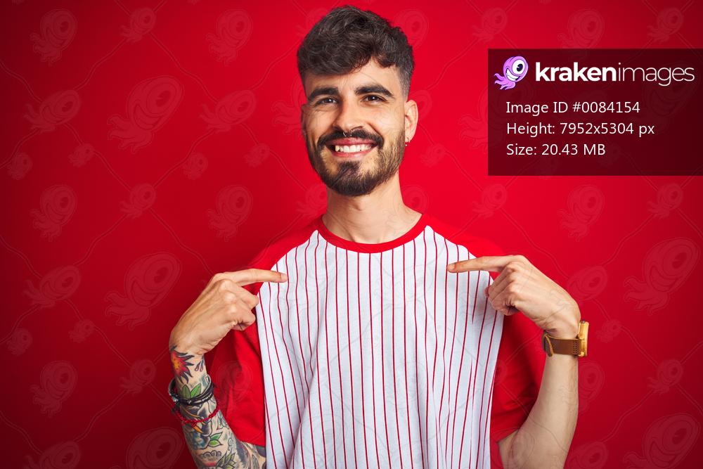 Young man with tattoo wearing striped t-shirt standing over isolated red background looking confident with smile on face, pointing oneself with fingers proud and happy.