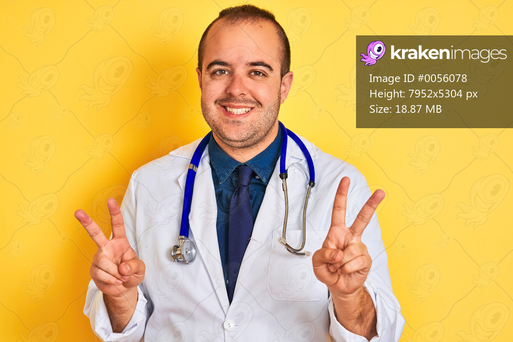 Young doctor man wearing coat and stethoscope standing over isolated yellow background smiling looking to the camera showing fingers doing victory sign. Number two.