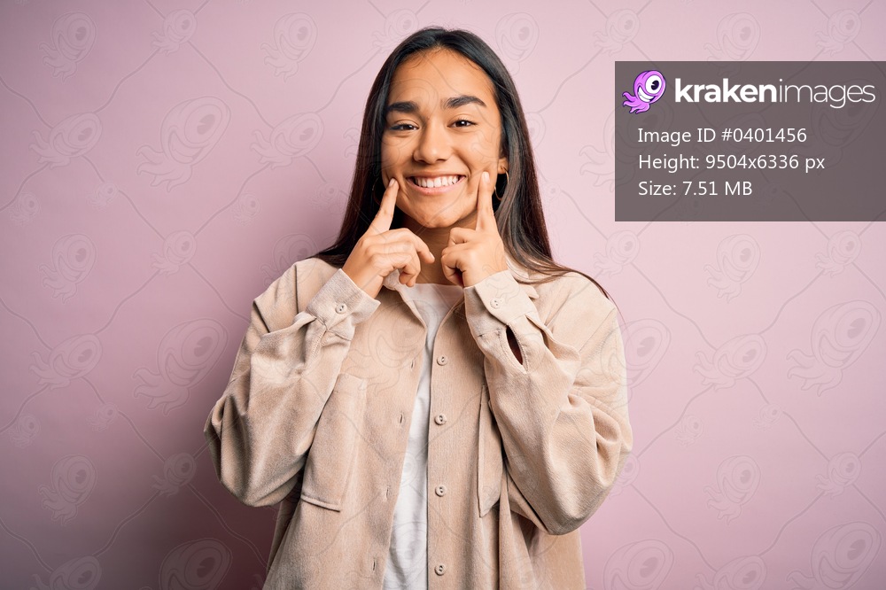 Young beautiful asian woman wearing casual shirt standing over pink background Smiling with open mouth, fingers pointing and forcing cheerful smile