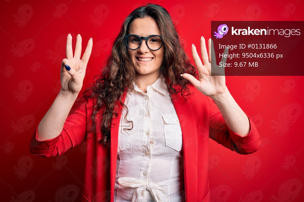 Young beautiful woman with curly hair wearing jacket and glasses over red background showing and pointing up with fingers number eight while smiling confident and happy.
