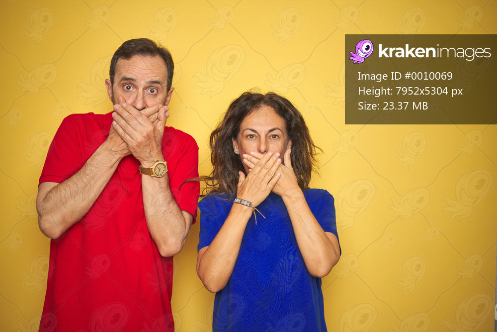 Beautiful middle age couple together standing over isolated yellow background shocked covering mouth with hands for mistake. Secret concept.