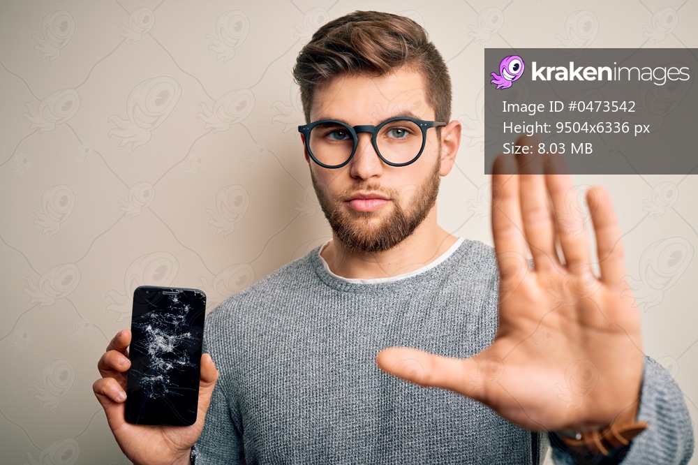 Young blond man with beard and blue eyes wearing glasses holding broken smartphone with open hand doing stop sign with serious and confident expression, defense gesture