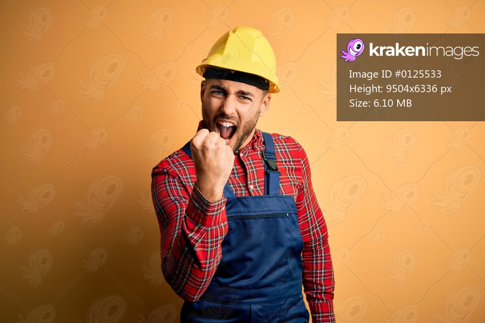 Young builder man wearing construction uniform and safety helmet over yellow isolated background angry and mad raising fist frustrated and furious while shouting with anger. Rage and aggressive concept.