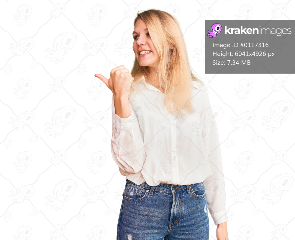 Young beautiful blonde woman wearing casual shirt smiling with happy face looking and pointing to the side with thumb up. 