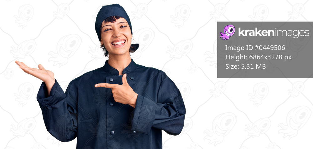 Young brunette woman with short hair wearing professional cook uniform amazed and smiling to the camera while presenting with hand and pointing with finger. 