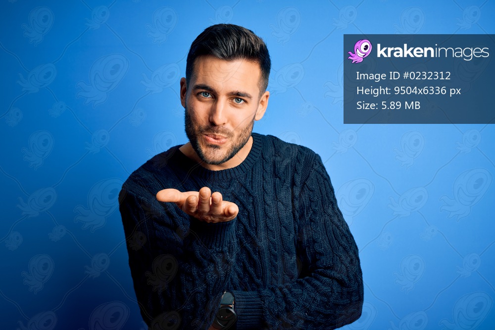 Young handsome man wearing casual sweater standing over isolated blue background looking at the camera blowing a kiss with hand on air being lovely and sexy. Love expression.