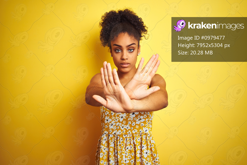 African american woman wearing casual floral dress standing over isolated yellow background Rejection expression crossing arms and palms doing negative sign, angry face