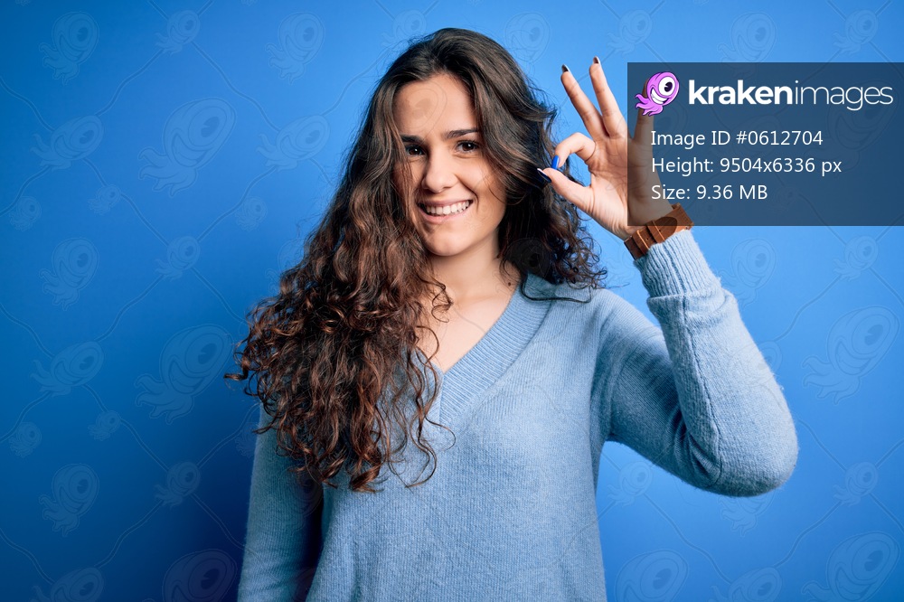 Young beautiful woman with curly hair wearing blue casual sweater over isolated background smiling positive doing ok sign with hand and fingers. Successful expression.