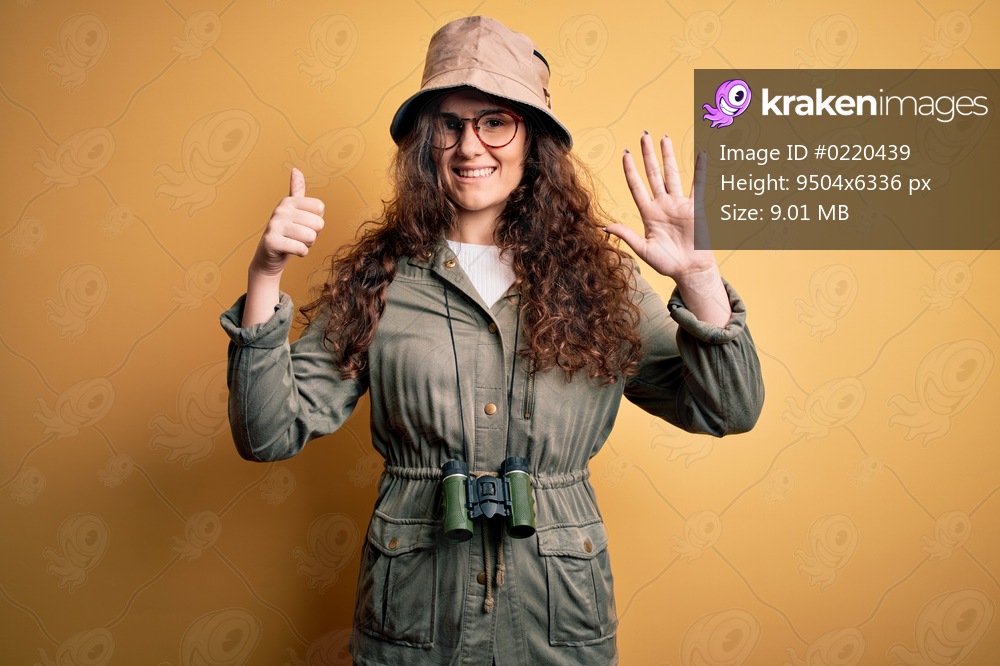 Young beautiful tourist woman on vacation wearing explorer hat and binoculars showing and pointing up with fingers number six while smiling confident and happy.