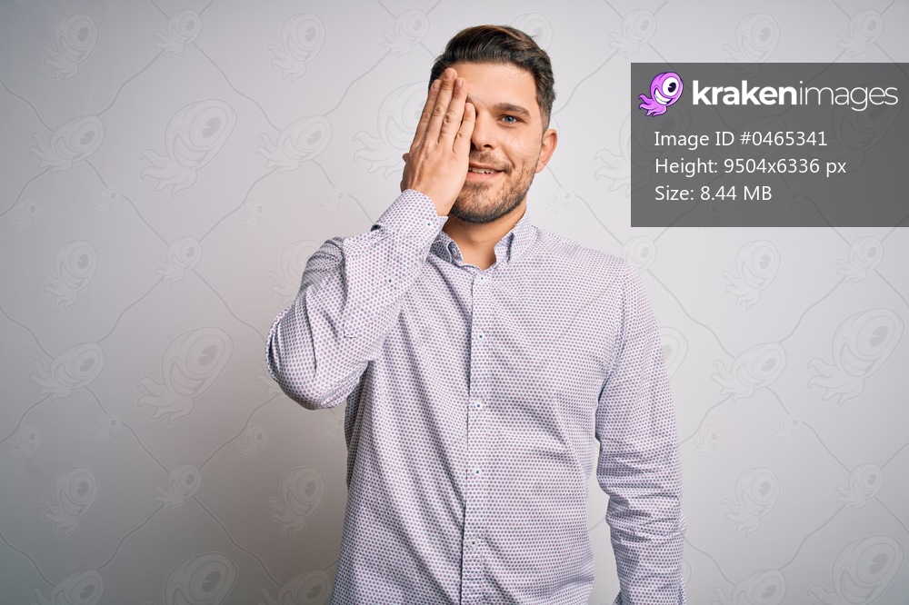 Young business man with blue eyes standing over isolated background covering one eye with hand, confident smile on face and surprise emotion.