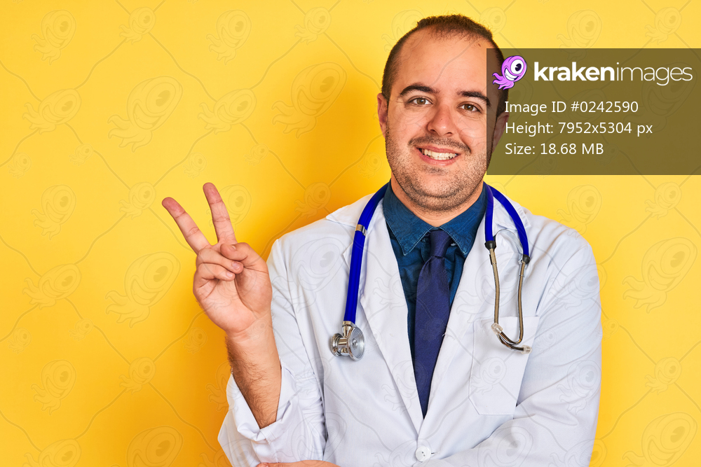 Young doctor man wearing coat and stethoscope standing over isolated yellow background smiling with happy face winking at the camera doing victory sign. Number two.