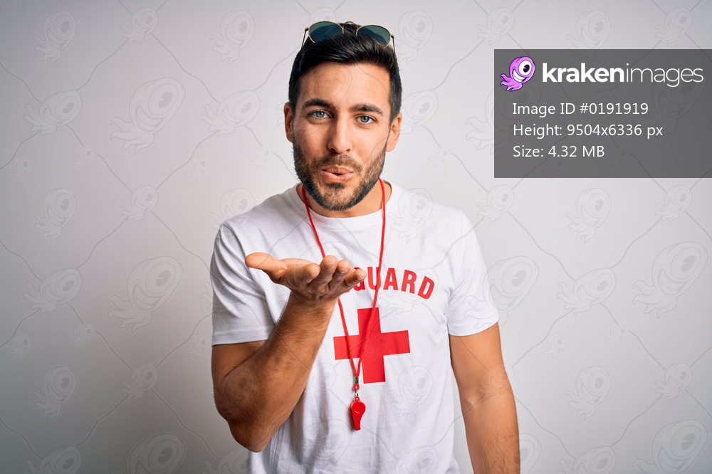 Young handsome lifeguard man with beard wearing t-shirt with red cross and whistle looking at the camera blowing a kiss with hand on air being lovely and sexy. Love expression.