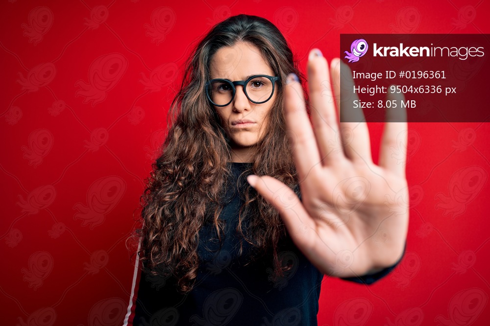 Young beautiful woman with curly hair wearing sweater and glasses over red background doing stop sing with palm of the hand. Warning expression with negative and serious gesture on the face.