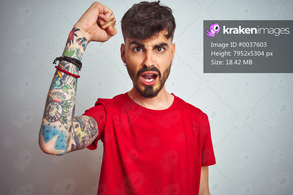 Young man with tattoo wearing red t-shirt standing over isolated white background angry and mad raising fist frustrated and furious while shouting with anger. Rage and aggressive concept.