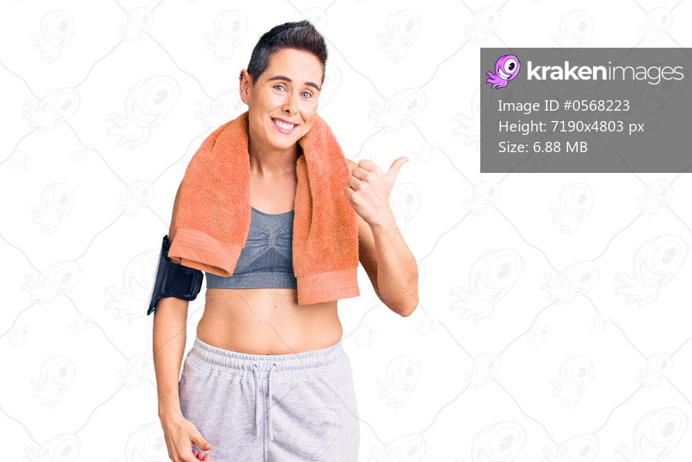 Young woman with short hair wearing sportswear and towel using smartphone smiling with happy face looking and pointing to the side with thumb up. 