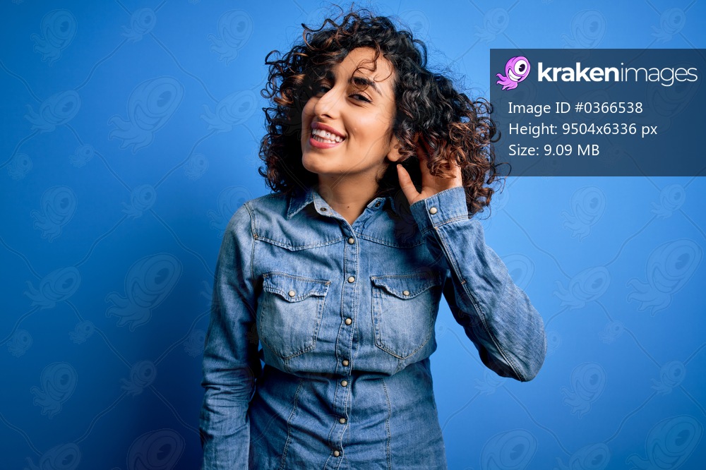 Young beautiful curly arab woman wearing casual denim shirt standing over blue background smiling with hand over ear listening an hearing to rumor or gossip. Deafness concept.