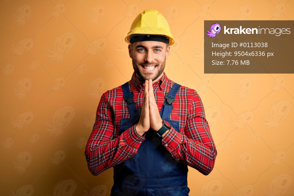 Young builder man wearing construction uniform and safety helmet over yellow isolated background praying with hands together asking for forgiveness smiling confident.