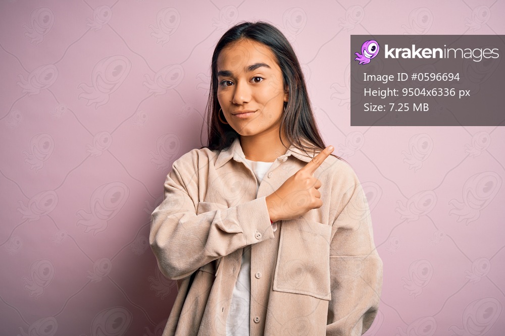 Young beautiful asian woman wearing casual shirt standing over pink background Pointing with hand finger to the side showing advertisement, serious and calm face