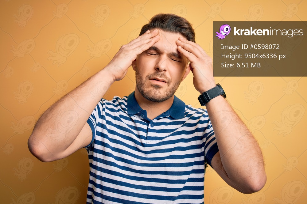Young man with blue eyes wearing casual striped t-shirt over yellow background suffering from headache desperate and stressed because pain and migraine. Hands on head.